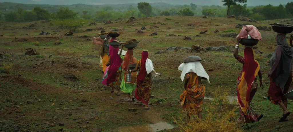 A group of women walking in a line through rural terrain carrying baskets on their heads | Co-Impact.