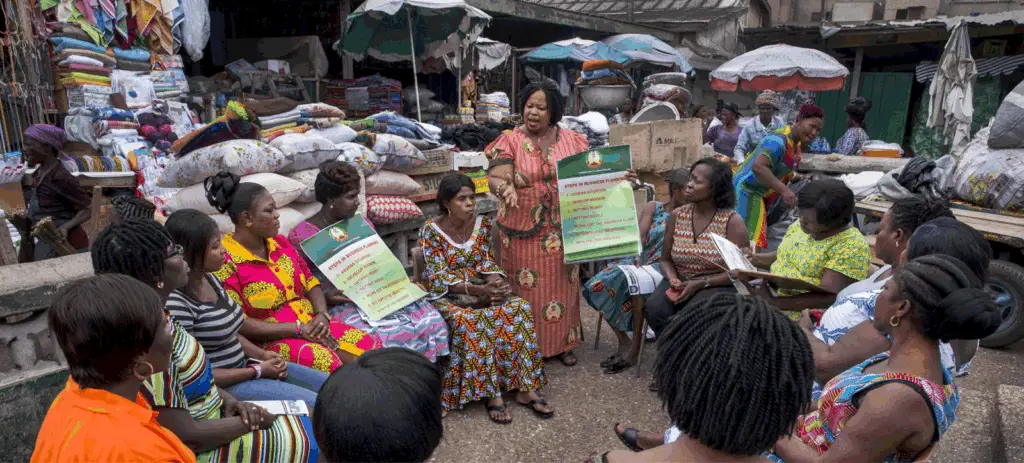 A woman leads a business planning workshop for market women in an outdoor setting surrounded by fabrics and goods, promoting just and inclusive systems change.
