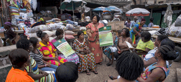 A woman leads a business planning workshop for market women in an outdoor setting surrounded by fabrics and goods, promoting just and inclusive systems change.
