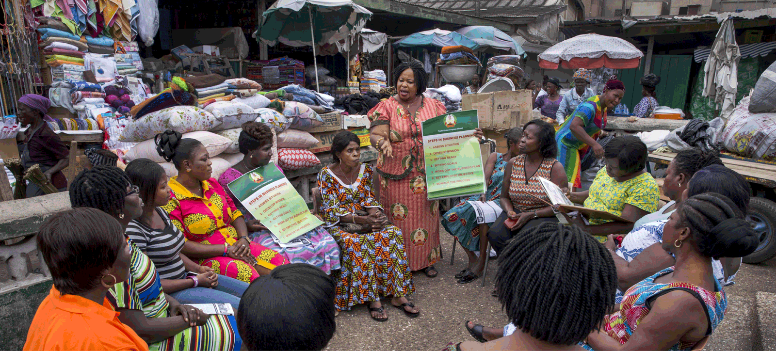 A woman leads a business planning workshop for market women in an outdoor setting surrounded by fabrics and goods, promoting just and inclusive systems change.