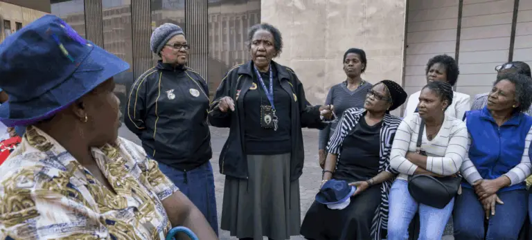An elder woman addresses a group of seated and standing women during a community gathering outdoors, discussing collective action and scaling local movements.