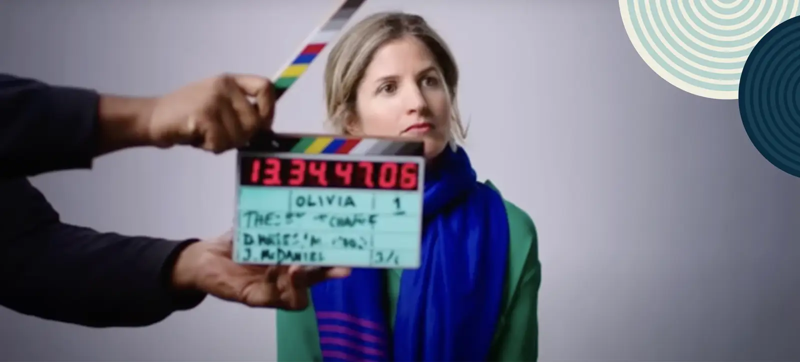 A woman sits in front of a camera during a film shoot, as a crew member holds a clapperboard reading “How to Change the World” and her name, Olivia.