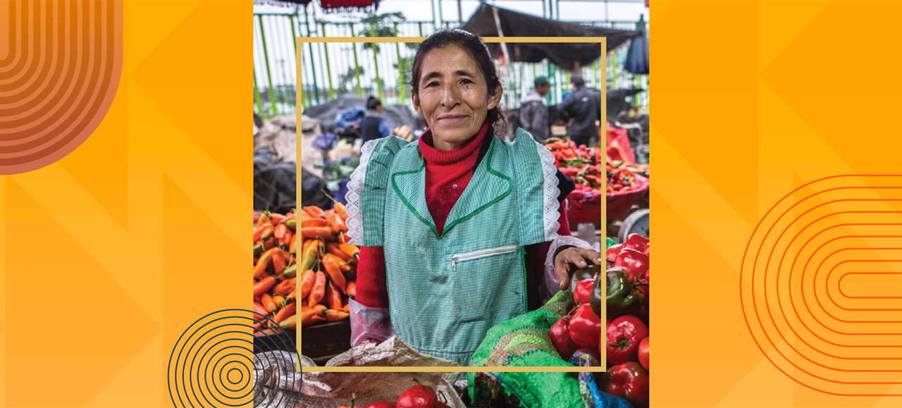 Woman vendor smiling at a vegetable market stall with chili peppers and bell peppers.