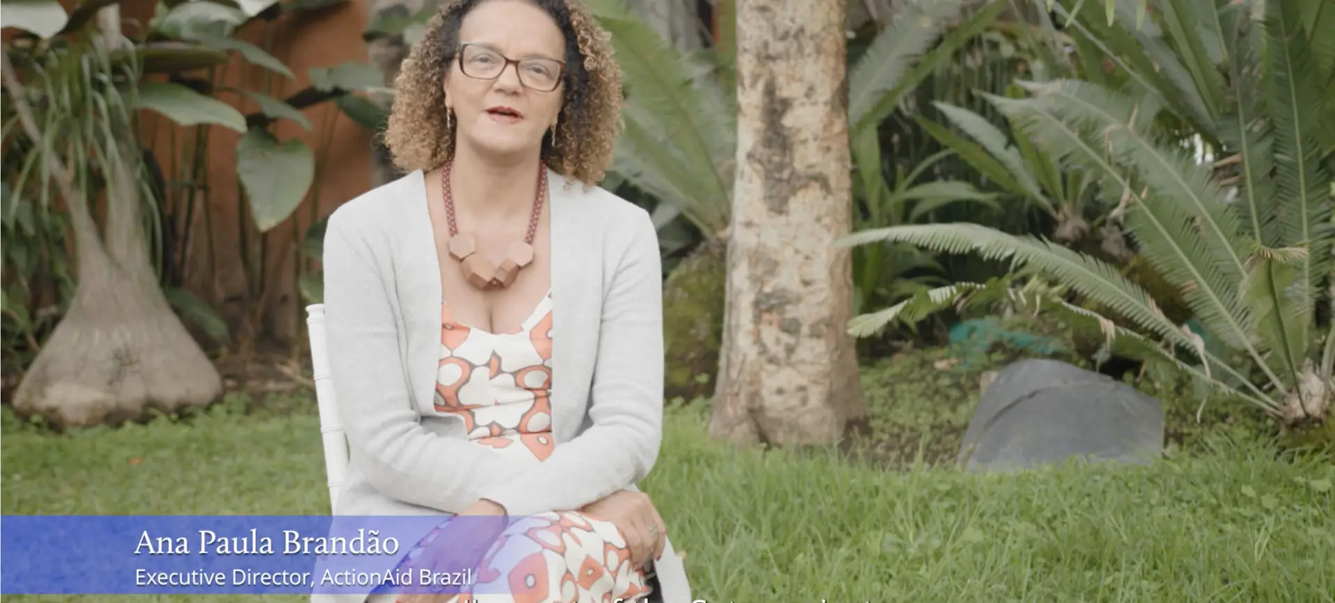 Ana Paula Brandão, a woman with curly hair and glasses wearing a white cardigan and patterned top, sitting outdoors in a tropical garden setting with palm trees and greenery in the background