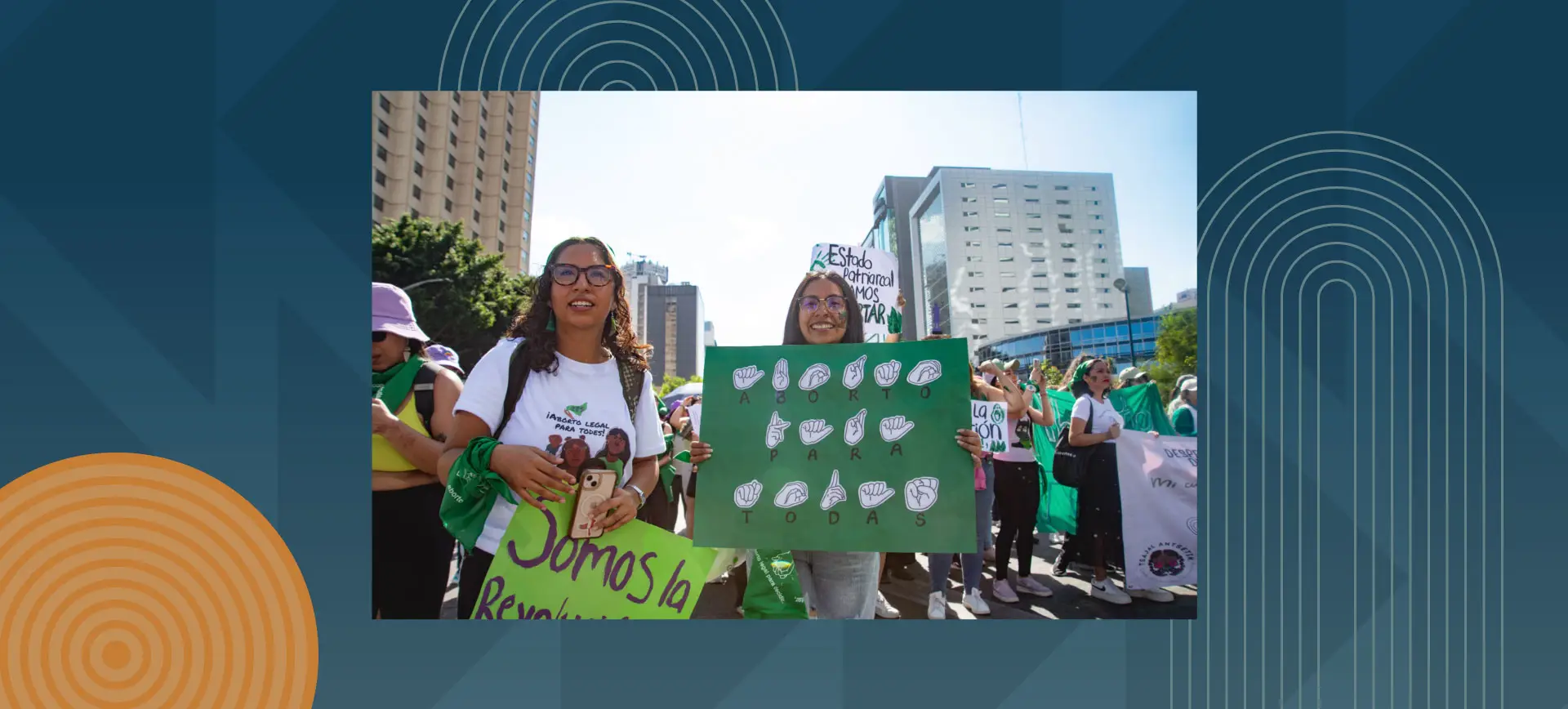 Two young women with glasses holding green protest signs at an outdoor demonstration in an urban setting, surrounded by other protesters wearing green and holding banners, with tall buildings visible in the background