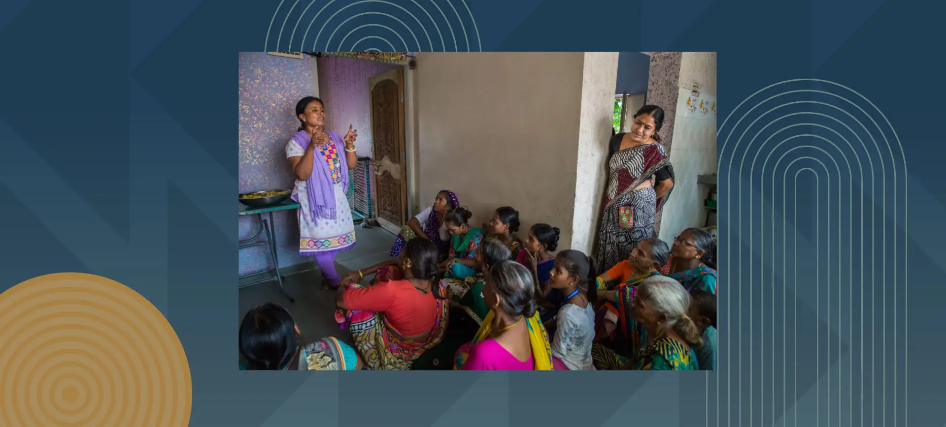 A woman stands and speaks to a group of women seated on the floor inside a home, during a community gathering in South Asia.