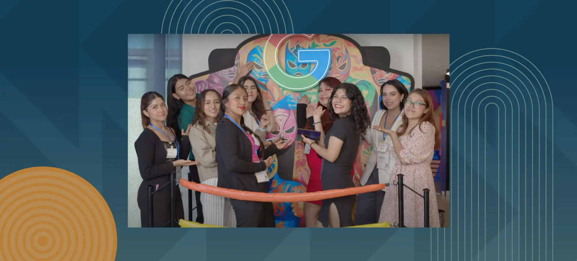 Group of young women posing together at an event, holding an orange ceremonial ribbon in front of a colorful wall with Google logo, all smiling and wearing identification badges