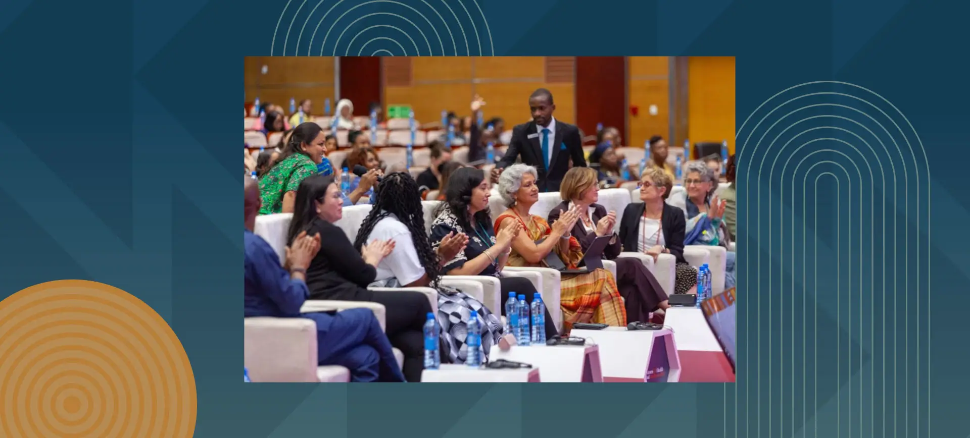 A diverse group of women and men sit in a large conference hall, smiling, applauding, and engaging in conversation during a formal gathering.