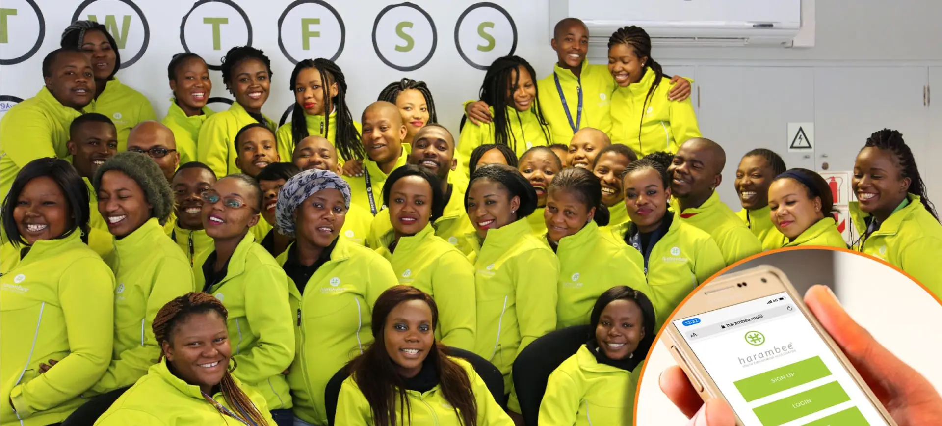 Large group of smiling African professionals in bright yellow uniforms posing together in office setting with company logo wall behind them, with mobile phone showing app interface in bottom right corner
