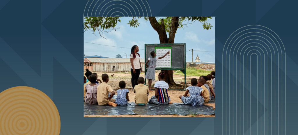 African children sitting on ground during outdoor classroom lesson with teacher and blackboard under tree