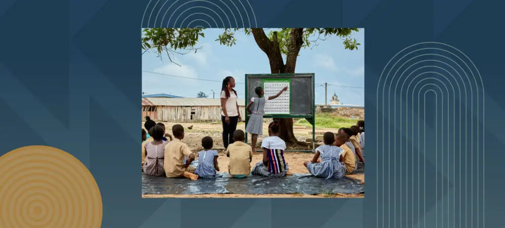 African children sitting on ground during outdoor classroom lesson with teacher and blackboard under tree