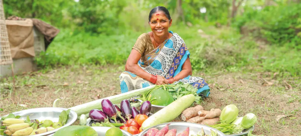 A woman smiling while sitting on the ground in front of a variety of fresh vegetables | Co-Impact.