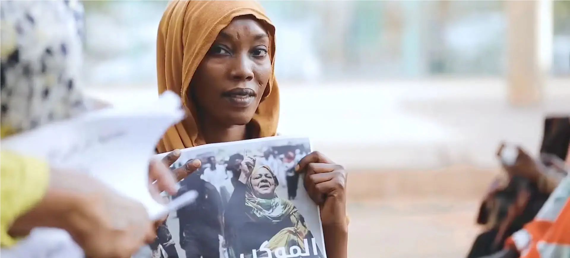 Woman in orange headscarf holding photograph during what appears to be a protest or demonstration