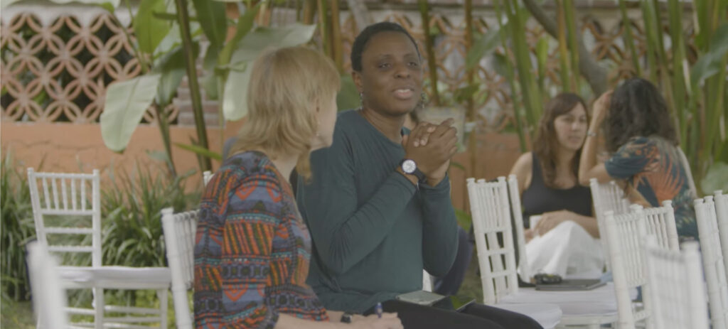 African woman speaking at outdoor event with microphone while blonde woman listens in garden setting